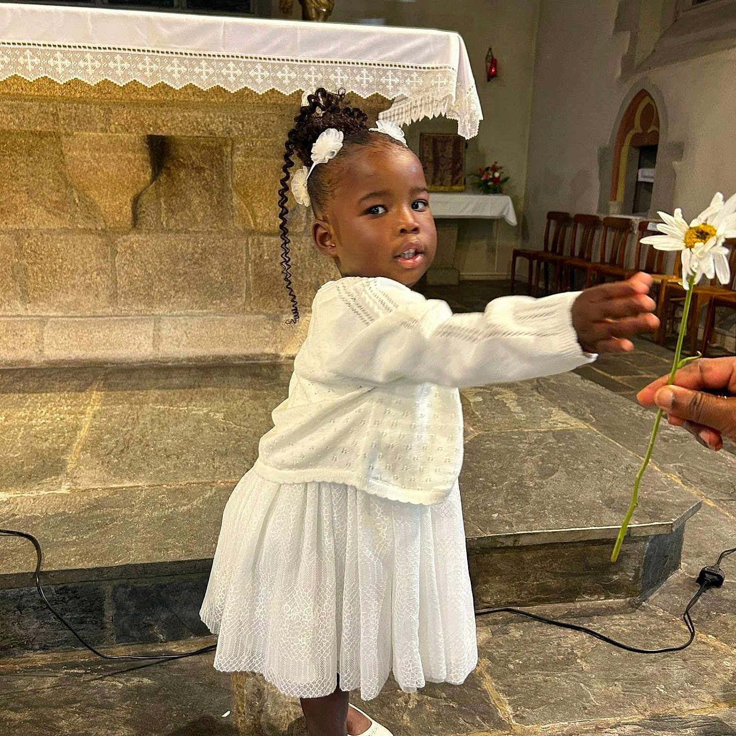 Gianna a rejoint le concours — aidez-le/la à gagner de superbes lots ! altar, bench, ceremony, child, church, curly_hair, daisy, decoration, flower, girl, hair_accessory, hand, indoor, offering, person, religious, shoes, stone_floor, white_dress, window