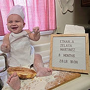 Ethan is registered to the contest to win money with this photo: baby, chef_hat, apron, flour, bread, kitchen, counter, smile, happy, infant, sign, wooden_board, cutting_board, cooking, messy, child, person, indoor, playful, cute