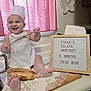 baby, chef_hat, apron, flour, bread, kitchen, counter, smile, happy, infant, sign, wooden_board, cutting_board, cooking, messy, child, person, indoor, playful, cute