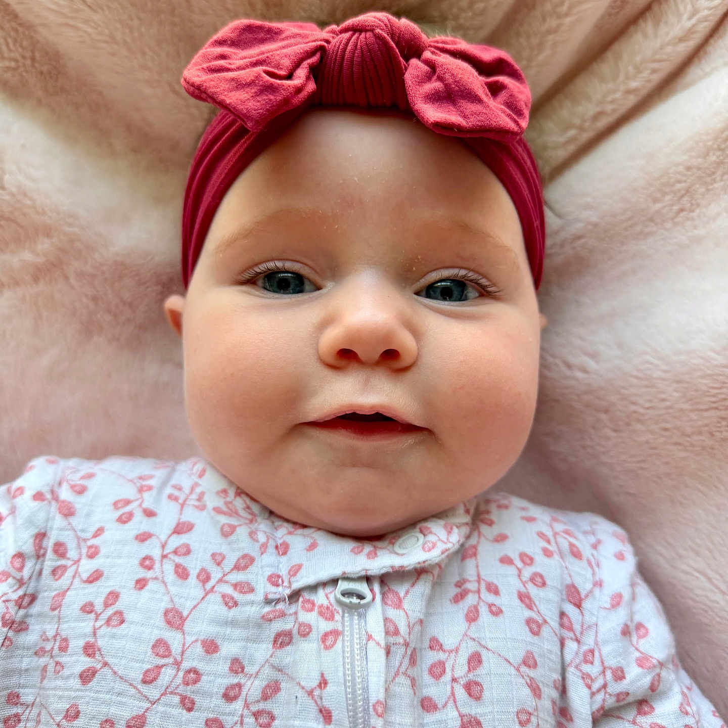 Emery is registered to the contest to win money with this photo: baby, infant, headband, red_bow, onesie, floral_pattern, pink_blanket, soft_texture, face, cheeks, eyes, portrait, cute, child, lying_down, indoors, closeup, expression, adorable, clothing