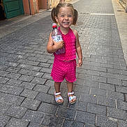 Joy a rejoint le concours — aidez-le/la à gagner de superbes lots ! child, smiling, girl, pink_clothing, water_bottle, pigtails, street, cobblestone, outdoor, footwear, sandals, happy, person, urban, daylight, sidewalk, standing, casual, young_child, people_in_background