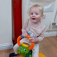 Ada-Rose is registered to the contest to win money with this photo: child, toddler, toy, turtle_toy, indoor, floor, wooden_floor, smiling, blonde_hair, long_sleeve_shirt, pants, socks, plastic_toy, handle, storage_pouches, wall, red_beam, electric_socket, radiator_pipe, happy