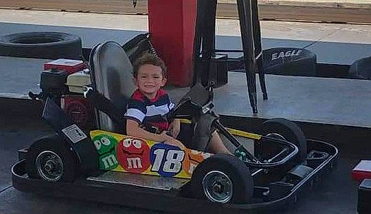 Kolby is registered to the contest to win money with this photo: automotive_design, automotive_exterior, automotive_tire, automotive_wheel_system, car, event, go_kart, helmet, joy, motor_vehicle, motorsport, person, race_track, racing, riding_toy, rim, smile, sports, tire, toddler