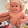 adult_arm, adult_hand, baby, cheeks, chef_hat, child, colorful_mat, diaper, eyes, face, furniture, holding, indoors, infant, playroom, portrait, smiling, tattoo, toys, window_blinds