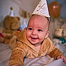 baby, birthday, birthday_hat, balloons, teddy_bear, smiling, happy, infant, child, portrait, close_up, indoor, celebration, party, brown_clothing, hooded_outfit, bed, soft_lighting, nursery, photography