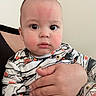 adult_hand, baby, caregiver, child, close_up, eyes, face, family, head, indoor, infant, looking_at_camera, onesie, patterned_clothing, portrait, protective, rosy_cheeks, safe, sitting, wide_eyed