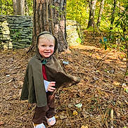 William joined the competition — help win amazing prizes! child, toddler, smile, hood, cloak, costume, forest, tree, tree_trunk, stone_wall, leaves, pine_needles, ground, outdoor, nature, sunlight, shoes, slippers, playful, portrait