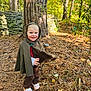 child, toddler, smile, hood, cloak, costume, forest, tree, tree_trunk, stone_wall, leaves, pine_needles, ground, outdoor, nature, sunlight, shoes, slippers, playful, portrait