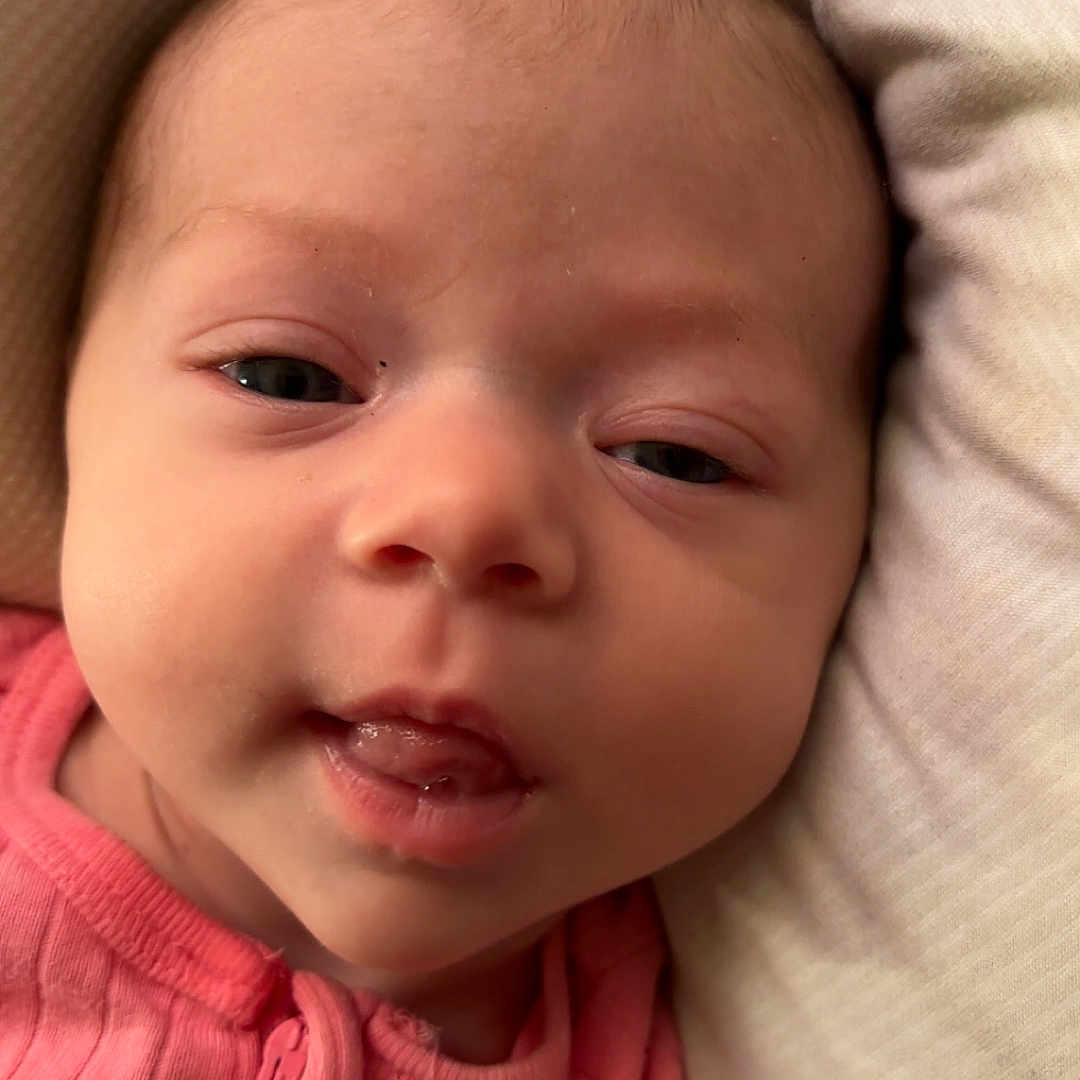 Isabel is registered to the contest to win money with this photo: baby, infant, face, pink_clothing, sleepy, tongue_out, close_up, cushion, soft_texture, newborn, child, portrait, cute, expression, indoor, person, young, skin, head, relaxed