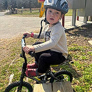Robert is registered to the contest to win money with this photo: child, helmet, bike, outdoor, park, caution_sign, grass, trees, sunlight, casual_clothing, red_shoes, pavement, wooden_bench, young_child, curious_expression, small_bike, nature, daylight, safety_gear, recreation
