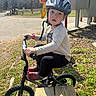 child, helmet, bike, outdoor, park, caution_sign, grass, trees, sunlight, casual_clothing, red_shoes, pavement, wooden_bench, young_child, curious_expression, small_bike, nature, daylight, safety_gear, recreation