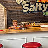 child, pizza, counter, restaurant, food, plate, sign, wood_wall, red_stool, bar_stool, interior, kid, table, snack, casual, young_child, indoor, curious, colorful, wooden_counter