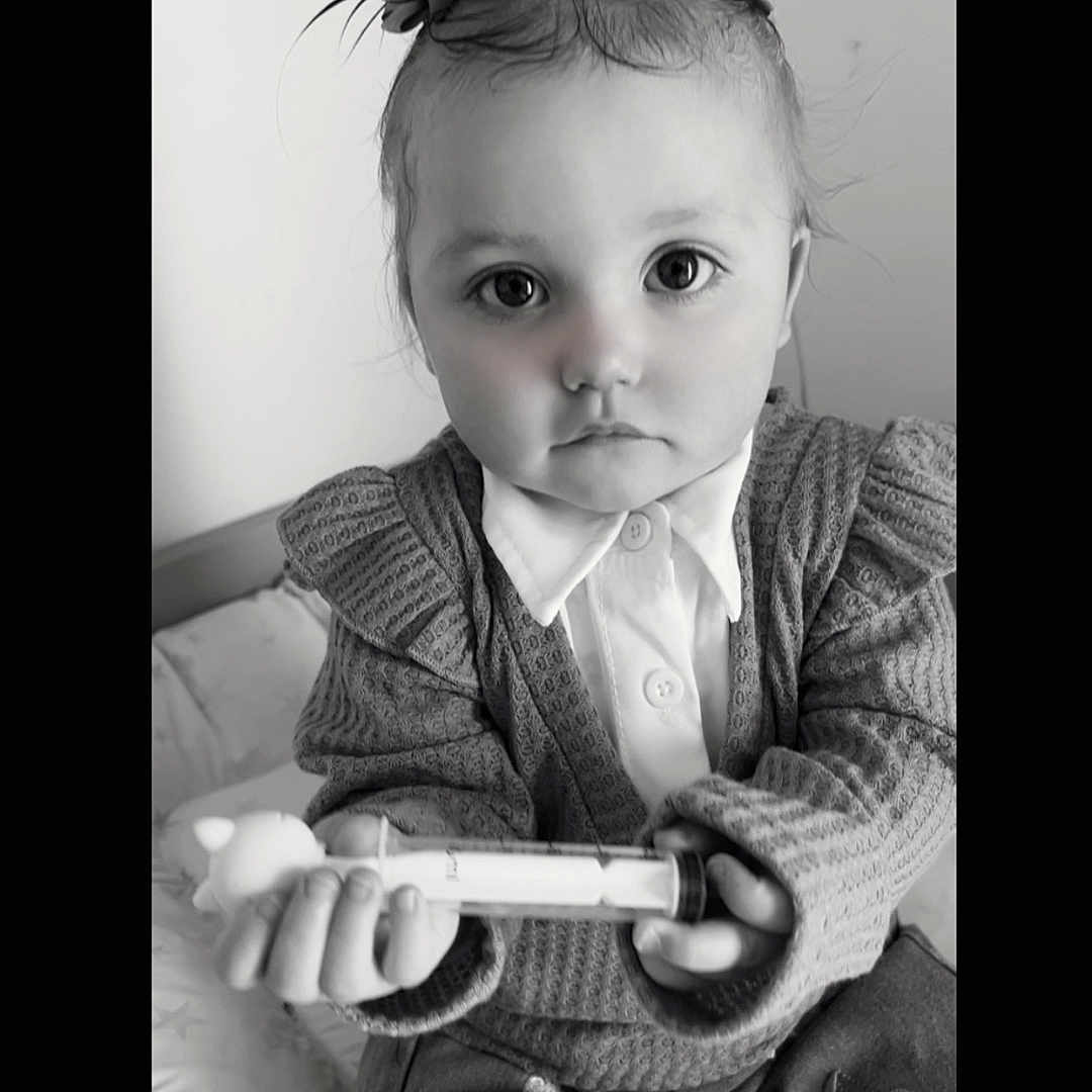 Leewena participe au concours pour gagner de l'argent avec cette photo : baby, bed, big_eyes, black_and_white, blanket, buttons, child, closeup, collared_shirt, cute, hair_bows, hands, indoor, portrait, serious_expression, sitting, sweater, syringe, toddler, toy