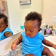 Aiden is registered to the contest to win money with this photo: baby, child, blue_outfit, glass_pitcher, mirror, reflection, bathroom, countertop, curious, sitting, infant, shorts, sleeveless, skin, hair, indoors, portrait, hand, looking_down, expression