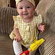 Persephone is registered to the contest to win money with this photo: baby, child, yellow_dress, headband, toy_gun, couch, indoor, smiling, sitting, cute, portrait, furniture, infant, happy, person, clothing, legs, feet, hand, face