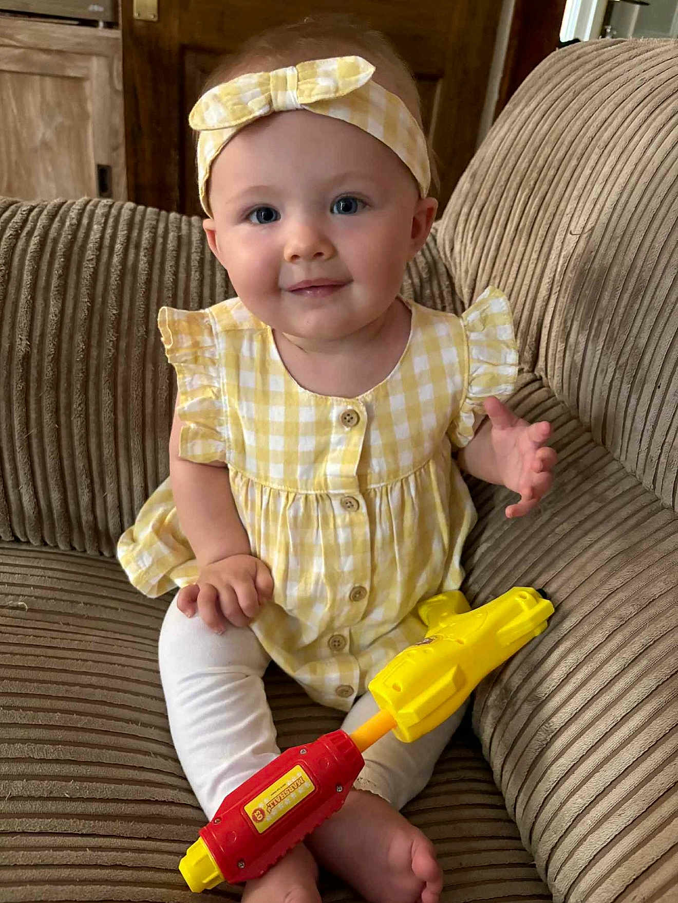 Persephone is registered to the contest to win money with this photo: baby, child, yellow_dress, headband, toy_gun, couch, indoor, smiling, sitting, cute, portrait, furniture, infant, happy, person, clothing, legs, feet, hand, face