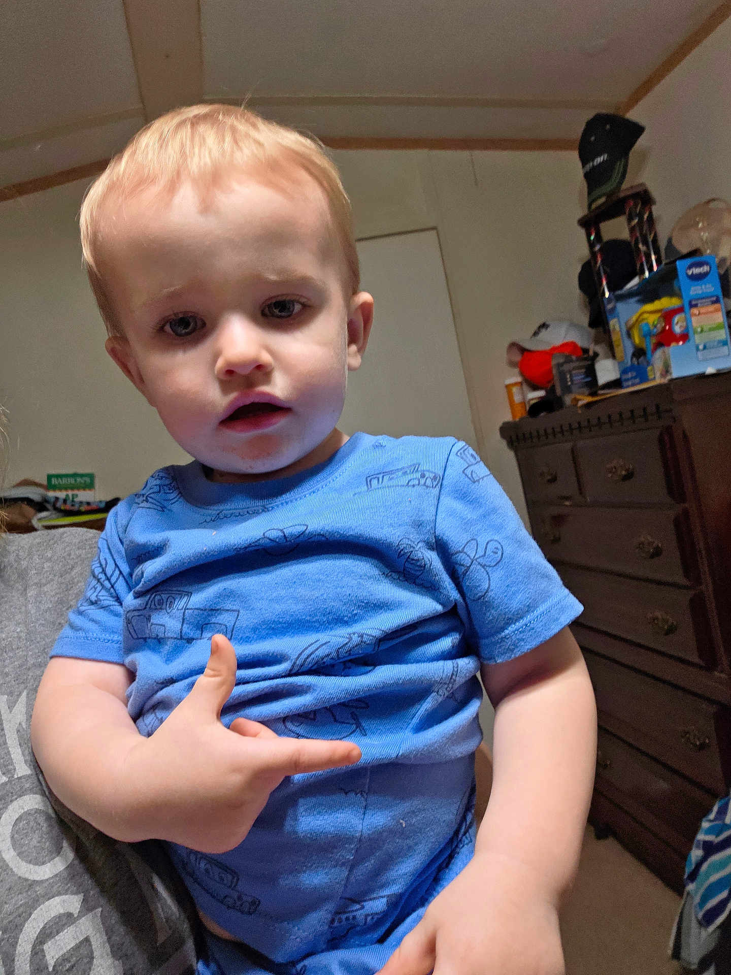 Sawyer is registered to the contest to win money with this photo: toddler, child, blue_shirt, indoor, person, furniture, dresser, clutter, toy_box, face, hand, light_hair, curious, casual, home, room, carpet, adult_hand, expression, portrait