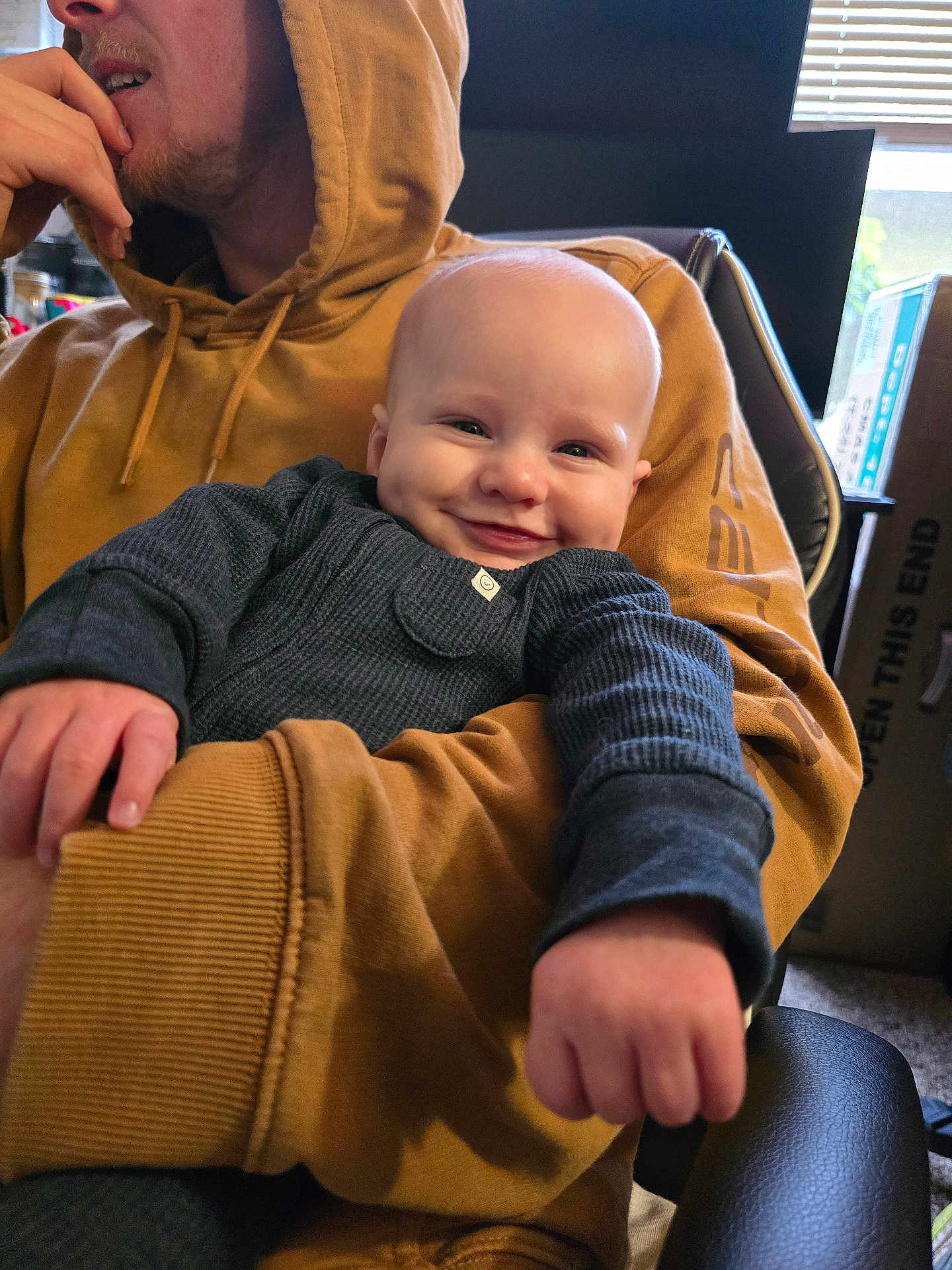 Ryker is registered to the contest to win money with this photo: baby, adult, smiling, blue_clothing, hoodie, indoor, window, natural_light, box, chair, hand, face, person, seated, cozy, casual, expression, portrait, holding, happy