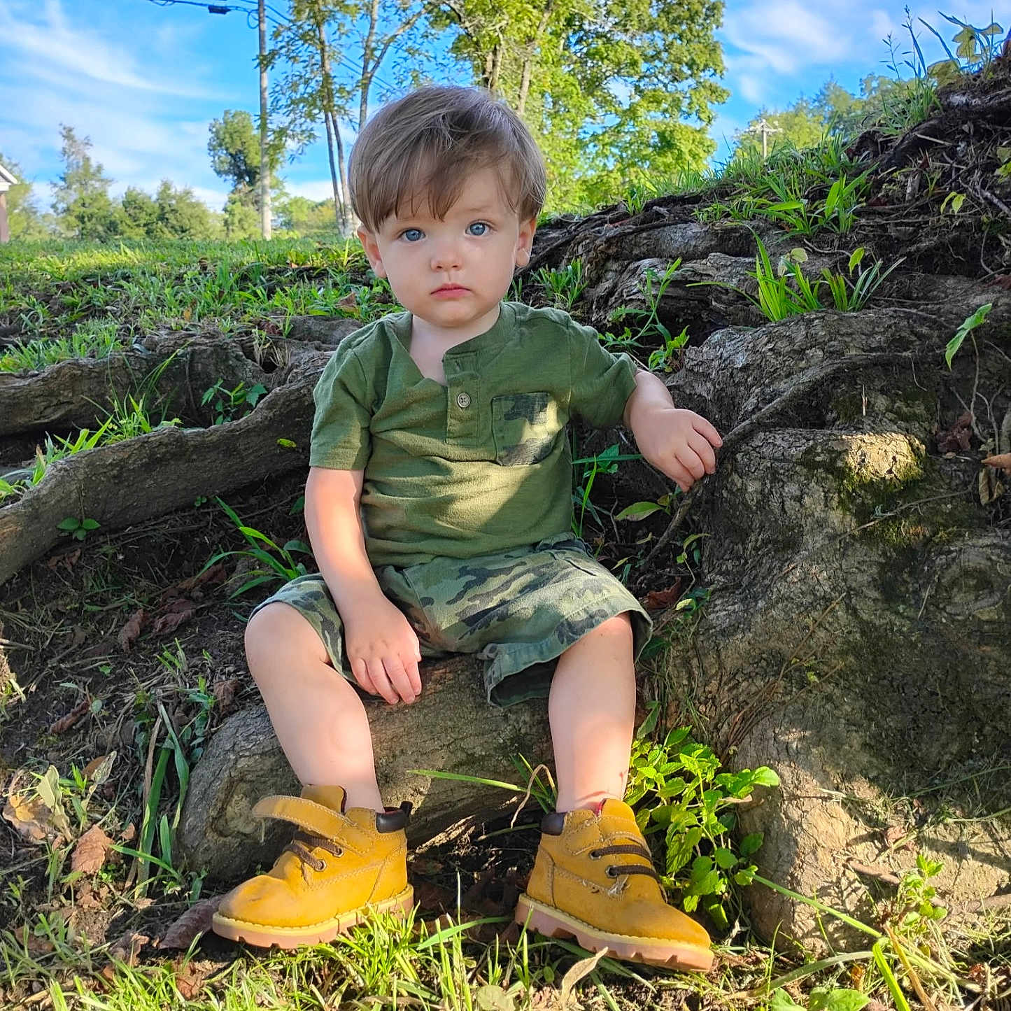 Odon joined the competition — help win amazing prizes! blue_sky, camo_shorts, casual_clothing, child, daylight, grass, green_shirt, nature, outdoor, person, portrait, serious_expression, shoes, sitting, sunlight, toddler, tree, tree_root, yellow_boots, young_child