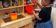 Riley is registered to the contest to win money with this photo: black_hair, black_shirt, blender_toy, blue_skirt, canned_food, child, flooring, focused, indoor, kitchen_sink, orange_pot, pink_bow, plastic_toys, play_food, pretend_play, sneakers, socks, toddler, toy_kitchen, wooden_furniture