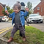 child, toddler, grass, curb, street, sidewalk, car, building, tree, clothing, boots, denim_jacket, hair_bun, smiling, outdoor, daytime, urban, residential, toy, happy