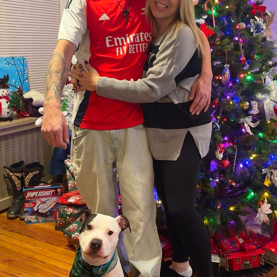 Yoshi is registered to the contest to win money with this photo: couple, dog, christmas_tree, holiday, indoor, hugging, smiling, decorations, lights, presents, wooden_floor, window, boots, tattoo, casual_clothing, sneakers, leggings, bandana, festive, home