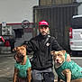 man, dog, indoor, kneeling, green_sweater, brown_dog, tan_dog, pet, animal, garage, van, casual_clothing, cap, beard, floor, equipment, calm, portrait, companionship, matching_outfits
