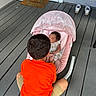 child, baby, baby_rocker, porch, wooden_floor, broom, welcome_mat, door, sneakers, orange_shirt, sleeping_baby, pink_rocker, mesh_cover, toddler, shorts, outdoor, floor, person, infant, shoes