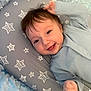 baby, infant, smiling, blue_clothing, pillow, star_pattern, blanket, lying_down, cute, indoors, face, happy, child, soft_texture, cozy, small_hand, young, head, closeup, person