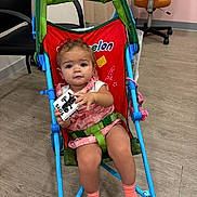 Ella is registered to the contest to win money with this photo: toddler, child, stroller, toy, indoor, chair, floor, socks, clothing, face, person, furniture, wood_floor, seat_belt, curly_hair, pink_outfit, blue_frame, playful, baby, expression