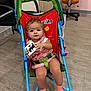 toddler, child, stroller, toy, indoor, chair, floor, socks, clothing, face, person, furniture, wood_floor, seat_belt, curly_hair, pink_outfit, blue_frame, playful, baby, expression