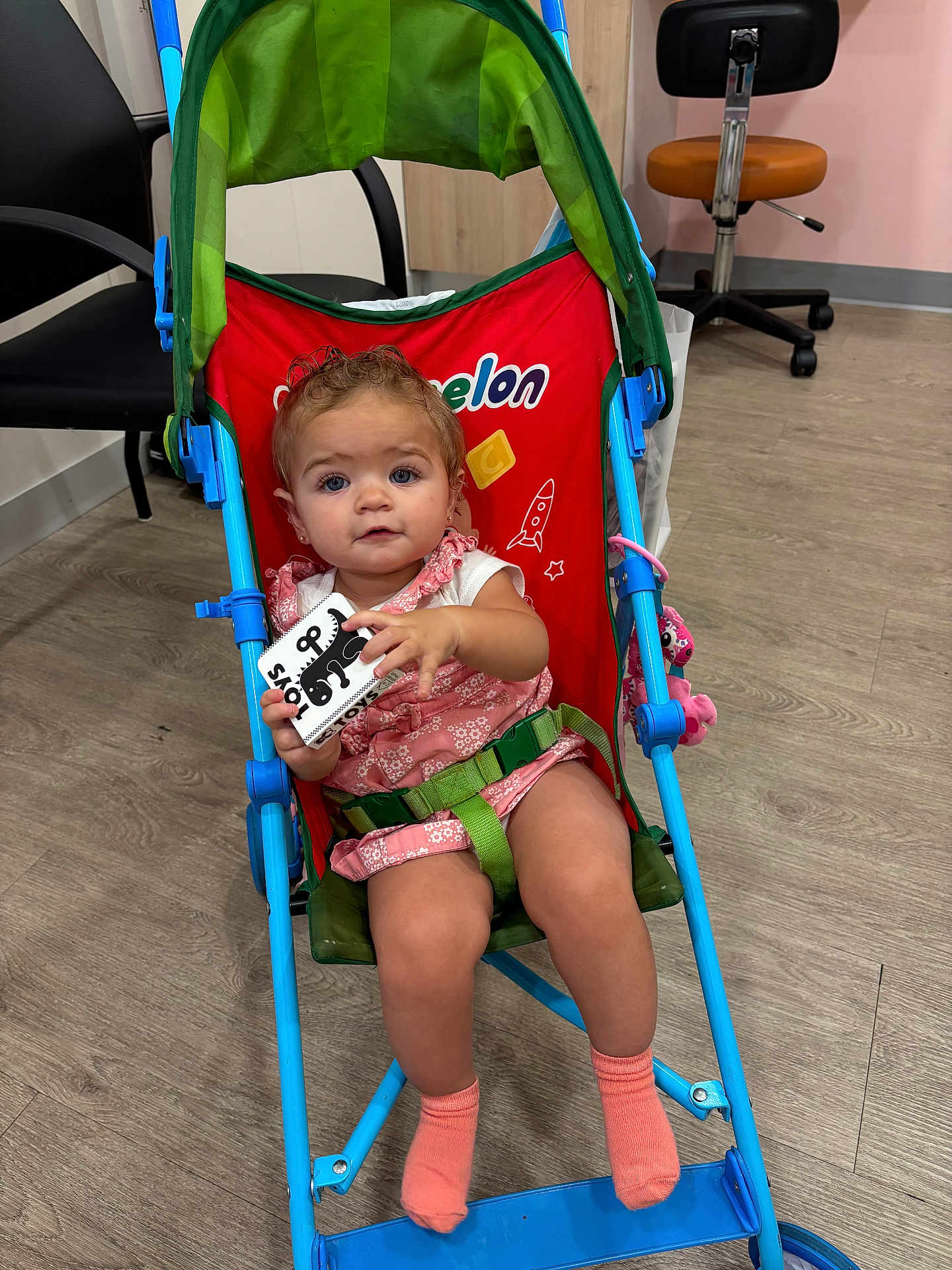Ella is registered to the contest to win money with this photo: toddler, child, stroller, toy, indoor, chair, floor, socks, clothing, face, person, furniture, wood_floor, seat_belt, curly_hair, pink_outfit, blue_frame, playful, baby, expression