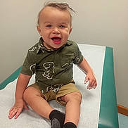 Gavin is registered to the contest to win money with this photo: baby, child, smiling, happy, portrait, sitting, dinosaur_shirt, shorts, socks, changing_table, indoor, home, wall, chair, green, white, clothing, face, hands, photography