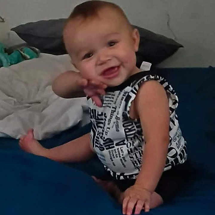 Gavin joined the competition — help win amazing prizes! baby, child, smiling, sitting, bed, bedroom, pillow, clothing, patterned_shirt, portrait, face, person, indoor, home, happy, playful, infant, toy, blue_bedding, casual