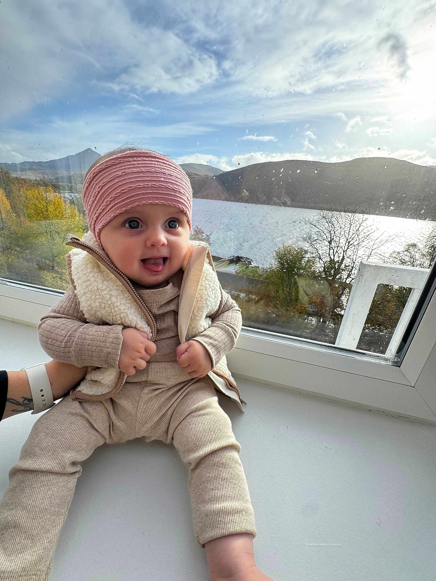 Oliwia joined the competition — help win amazing prizes! baby, child, window, lake, mountains, cloudy_sky, headband, vest, beige_clothing, tongue_out, blue_eyes, raindrops, nature, outdoor_view, smiling, sitting, cozy, hand, , , person