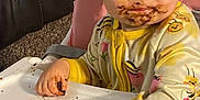 Jazalynn is registered to the contest to win money with this photo: toddler, child, high_chair, messy, chocolate, food, face, hand, yellow_clothing, onesie, bee_pattern, indoor, carpet, furniture, pink_chair, curious, cute, baby, portrait, eating