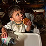 toddler, child, high_chair, sandwich, cup, striped_shirt, food, dining_room, wooden_chair, table, messy, curious, indoor, person, face, hand, drink, seat, furniture, floor