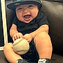 baby, smiling, baseball, cap, sneakers, black_shirt, denim_shorts, leather_couch, child, happy, indoor, person, cute, sport, toy, feet, seated, joyful, playful, face