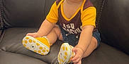 Carlos is registered to the contest to win money with this photo: baby, smiling, child, sitting, leather_couch, yellow_shirt, polka_dot_shoes, happy, indoors, cute, short_hair, denim_shorts, small_shoes, infant, cheerful, person, portrait, seated, couch, bright