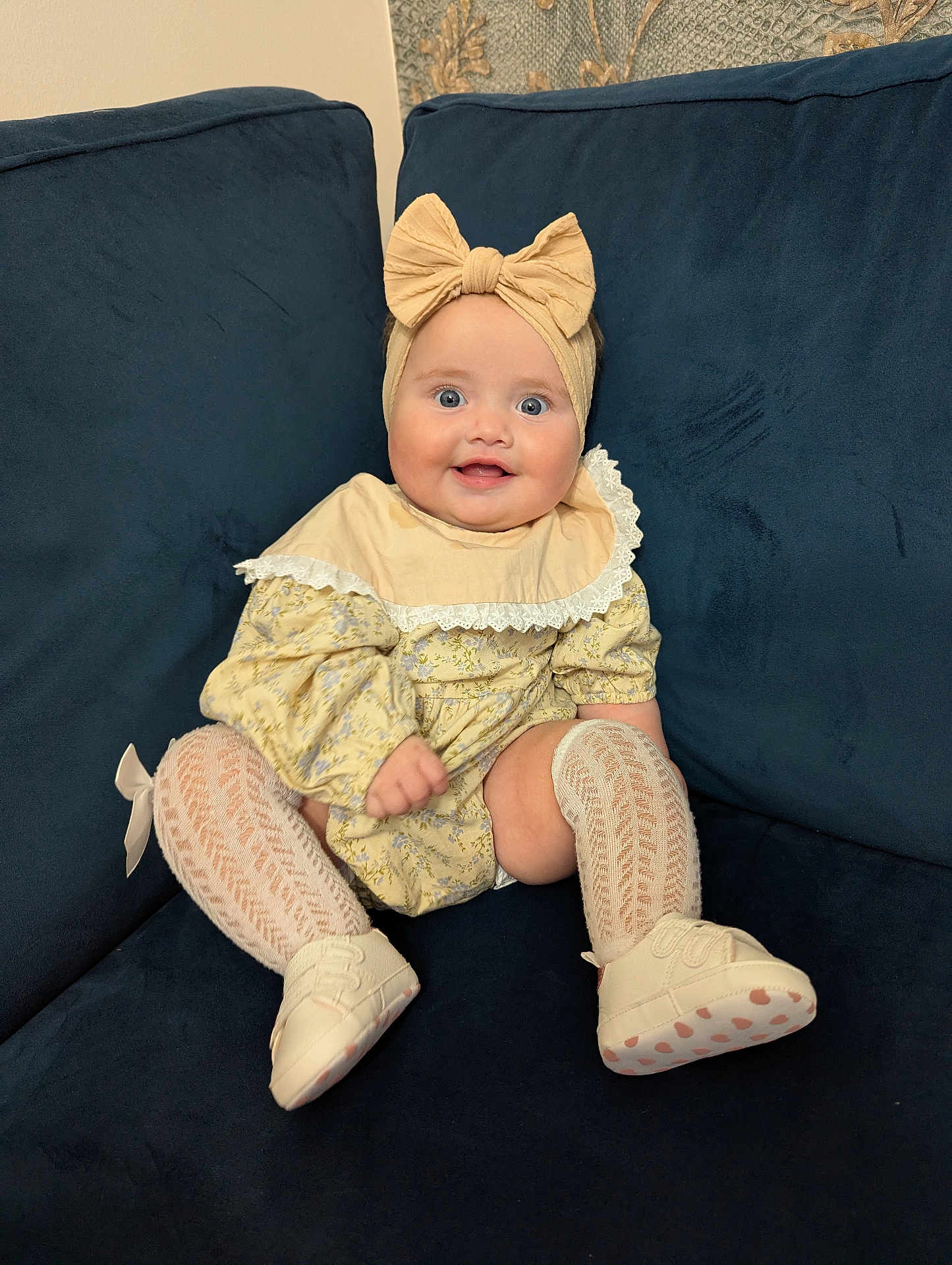 Evelyn joined the competition — help win amazing prizes! baby, infant, child, smiling, headband, bow, yellow_dress, sitting, couch, blue_couch, white_shoes, knit_socks, happy, portrait, indoor, cute, fashion, person, young_child, comfortable