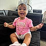 baby, child, smiling, pink_tshirt, white_bow, gold_chains, sneakers, sitting, couch, indoor, happy, headband, shorts, person, accessories, window, blinds, carpet, jewelry, cute