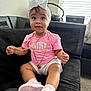 baby, child, girl, sitting, pink_clothing, headband, bow, gold_chain, sneakers, couch, black_couch, carpet, window, natural_light, indoor, curious, expression, person, shorts, footwear
