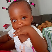 Elidja participe au concours pour gagner de l'argent avec cette photo : baby, child, infant, cute, hair_ties, puffs, hand_in_mouth, white_shirt, indoor, sofa, tablecloth, striped, earring, curious, close_up, person, home, floor, toy_clip, brown_skin