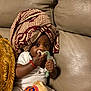 adorable, baby, blanket, child, clothing, comfort, couch, cute, headwrap, holding, indoor, infant, leather, pacifier, patterned_fabric, person, plush_toy, relaxed, sitting, toy