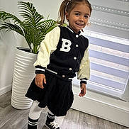 Mia participe au concours pour gagner de l'argent avec cette photo : child, girl, pigtails, smiling, indoor, plant, window, blinds, floor, sneakers, socks, skirt, jacket, letter_b, pot, greenery, happy, standing, casual_clothing, portrait