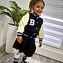 blinds, casual_clothing, child, floor, girl, greenery, happy, indoor, jacket, letter_b, pigtails, plant, portrait, pot, skirt, smiling, sneakers, socks, standing, window
