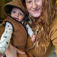 Elijah James joined the competition — help win amazing prizes! baby, infant, adult, woman, parent_child, smile, hooded_vest, long_hair, curly_hair, freckles, septum_piercing, indoor, cozy, brown_tones, holding, closeup, portrait, casual_clothing, wood_paneling, happy