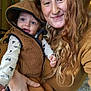 baby, infant, adult, woman, parent_child, smile, hooded_vest, long_hair, curly_hair, freckles, septum_piercing, indoor, cozy, brown_tones, holding, closeup, portrait, casual_clothing, wood_paneling, happy