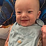baby, smile, bib, pillow, hand, tattoo, infant, face, clothing, pattern, blue, gold, couch, happy, skin, eyes, mouth, person, indoor, cute