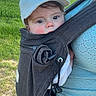 baby, child, carrier, white_cap, outdoor, green_field, blue_sky, adult, clothing, face, person, nature, grass, sunlight, portrait, young_child, holding, casual_wear, comfort, smiling